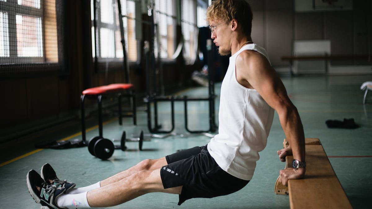 Side view of muscular sportsman in sportswear and glasses doing tricep dips exercises in gym